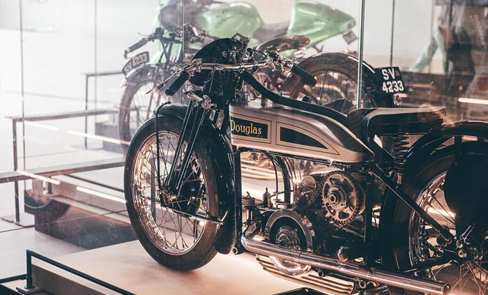 A silver Douglas motorbike in a display case in the foreground and a green motorbike in a display case in the background.